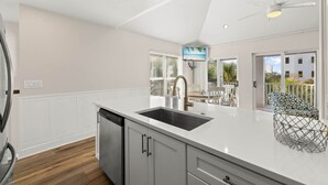 Een moderne keuken met een wit aanrecht, een roestvrijstalen spoelbak en een vaatwasser. Er is een plafondventilator, een raam met zonwering en uitzicht op buiten.