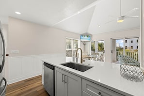 A modern kitchen with a white countertop, stainless steel sink, and a dishwasher. There is a ceiling fan, a window with blinds, and a view of the outside. - A Perfect Summer East (Walton County)