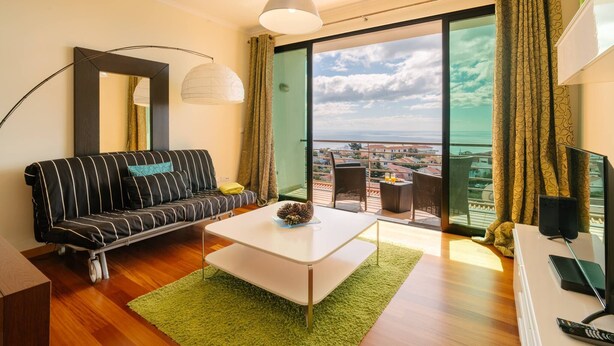 A modern living room with a sofa, a coffee table, a TV, and a balcony with a city view. - Ponta do Sol Apartment | Three-Bedroom (Ponta do Sol)