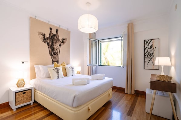 A bedroom with a large bed, a bedside table with a lamp, a window with curtains, and a wall art piece.