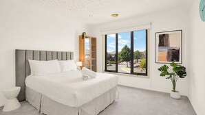 Ein Schlafzimmer mit Bett, einem Fenster mit Blick, einer Pflanze und einem Bild an der Wand.