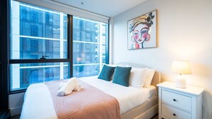 Ein Hotelzimmer mit großem Fenster, einem Bett mit Kissen, einem Nachttisch mit Lampe und einem Bild an der Wand.