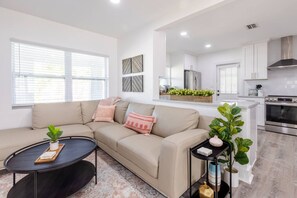 A modern living room with a beige sofa, a round coffee table, and a kitchen area with white cabinets and stainless steel appliances. - Walkable Tampa Bungalow Close to Top Spots (Tampa)