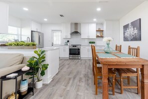 A modern kitchen with white cabinets, a wooden dining table, and a large window. - Walkable Tampa Bungalow Close to Top Spots (Tampa)