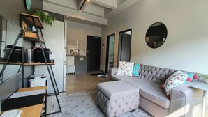 A modern living room with a grey sectional sofa, a small side table, a ladder with items on it, a mirror on the wall, and a door leading to a kitchen. - 15 Sunny 1-bedroom Apartment (Johannesburg)