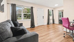 A spacious room with a grey sofa, a pink office chair, and a large window with curtains.