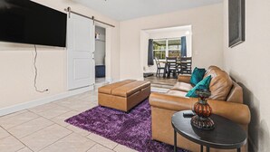 A living room with a sofa, ottoman, and a table with a lamp. There's a TV mounted on the wall and a ceiling fan.