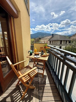 Une terrasse en bois avec une table et des chaises, une porte vitrée et une vue sur les montagnes et les bâtiments.
