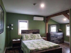 A bedroom with a wooden headboard, a bed with a floral bedspread, a nightstand, a dresser, and a floor lamp. - Cozy, tranquil waterfront retreat with a stunning oceanfront view in the pines. (Shelburne)