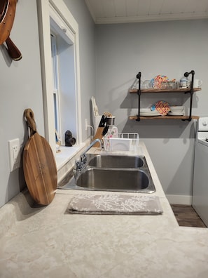 A kitchen with a double sink, wooden cutting board, and a shelf with dishes and glasses. - Cozy, tranquil waterfront retreat with a stunning oceanfront view in the pines. (Shelburne)
