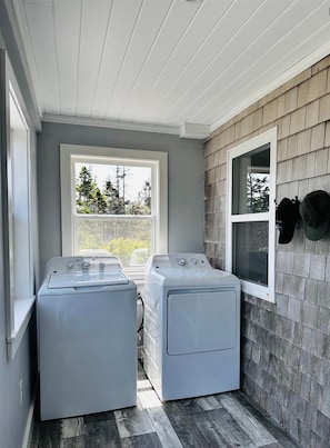 A laundry room with two front-loading washing machines, a window with a view of trees, and a wall-mounted hat rack. - Cozy, tranquil waterfront retreat with a stunning oceanfront view in the pines. (Shelburne)