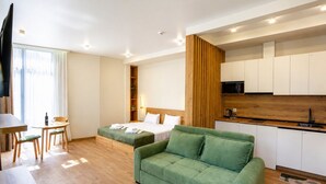 Une chambre d’hôtel moderne avec un plancher en bois, un canapé vert, un lit avec du linge de maison blanc, une petite table à manger avec des chaises, un coin cuisine et une grande fenêtre avec des rideaux.