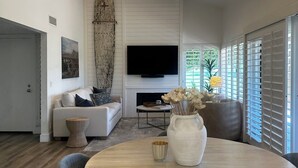 Una sala de estar moderna con una mesa de comedor redonda, un sofá, un televisor y un ventanal con persianas.