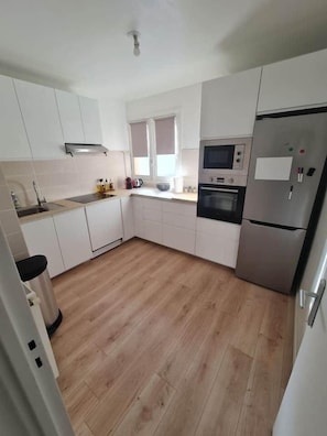 Eine kleine Küche mit weißen Schränken, einem Edelstahl-Kühlschrank, einer Mikrowelle und einem Fenster mit Jalousien.