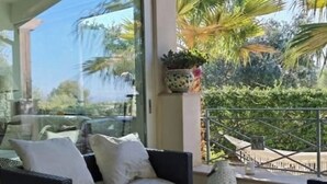 Un balcon avec un canapé en osier et des coussins, une table en verre et une plante en pot.