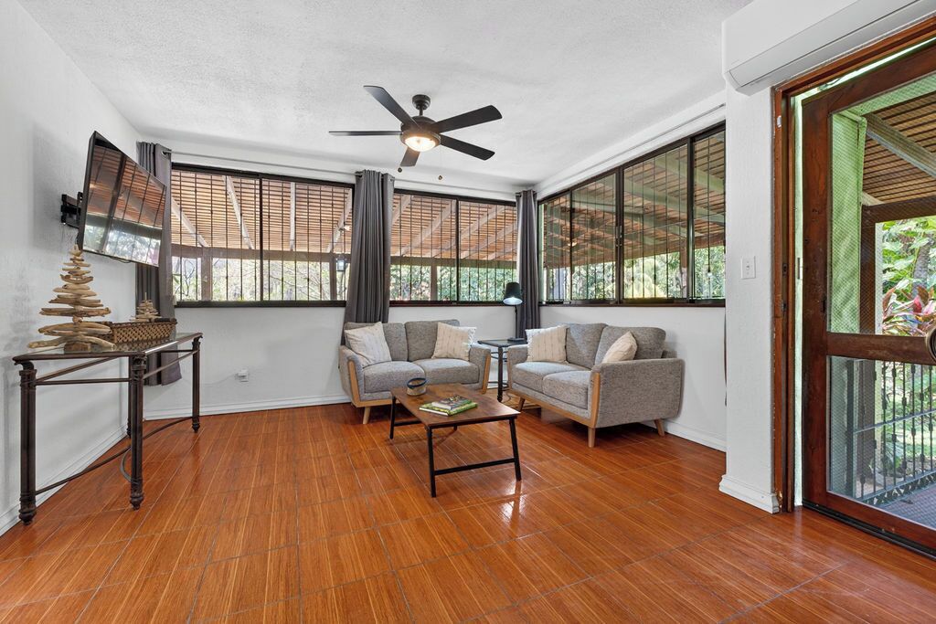 A living room with a wooden floor, two grey sofas, a coffee table, a ceiling fan, and large windows with blinds.