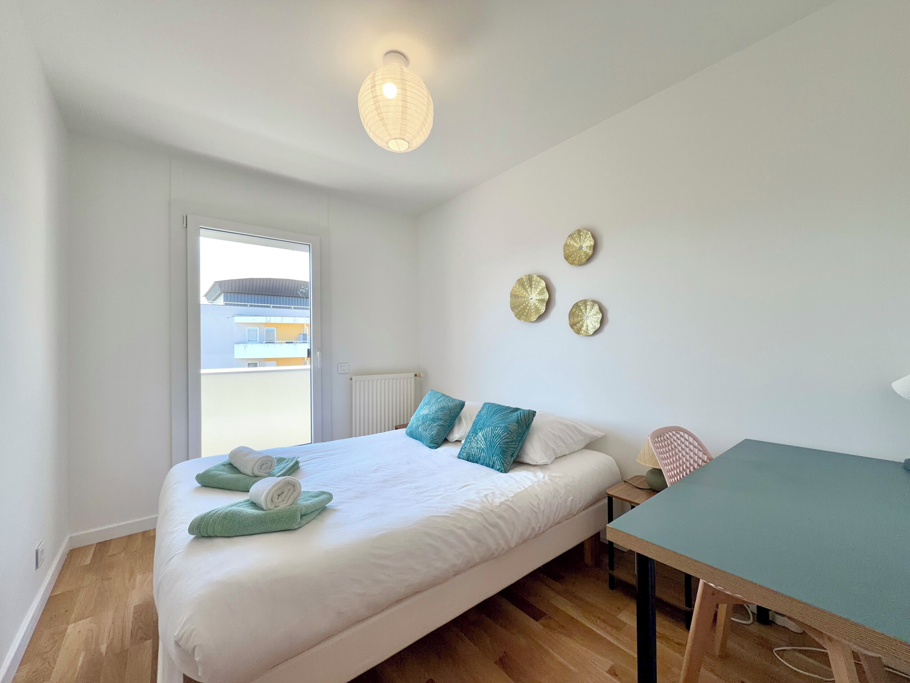 A bedroom with a bed, a table, a lamp, and a view through a window.