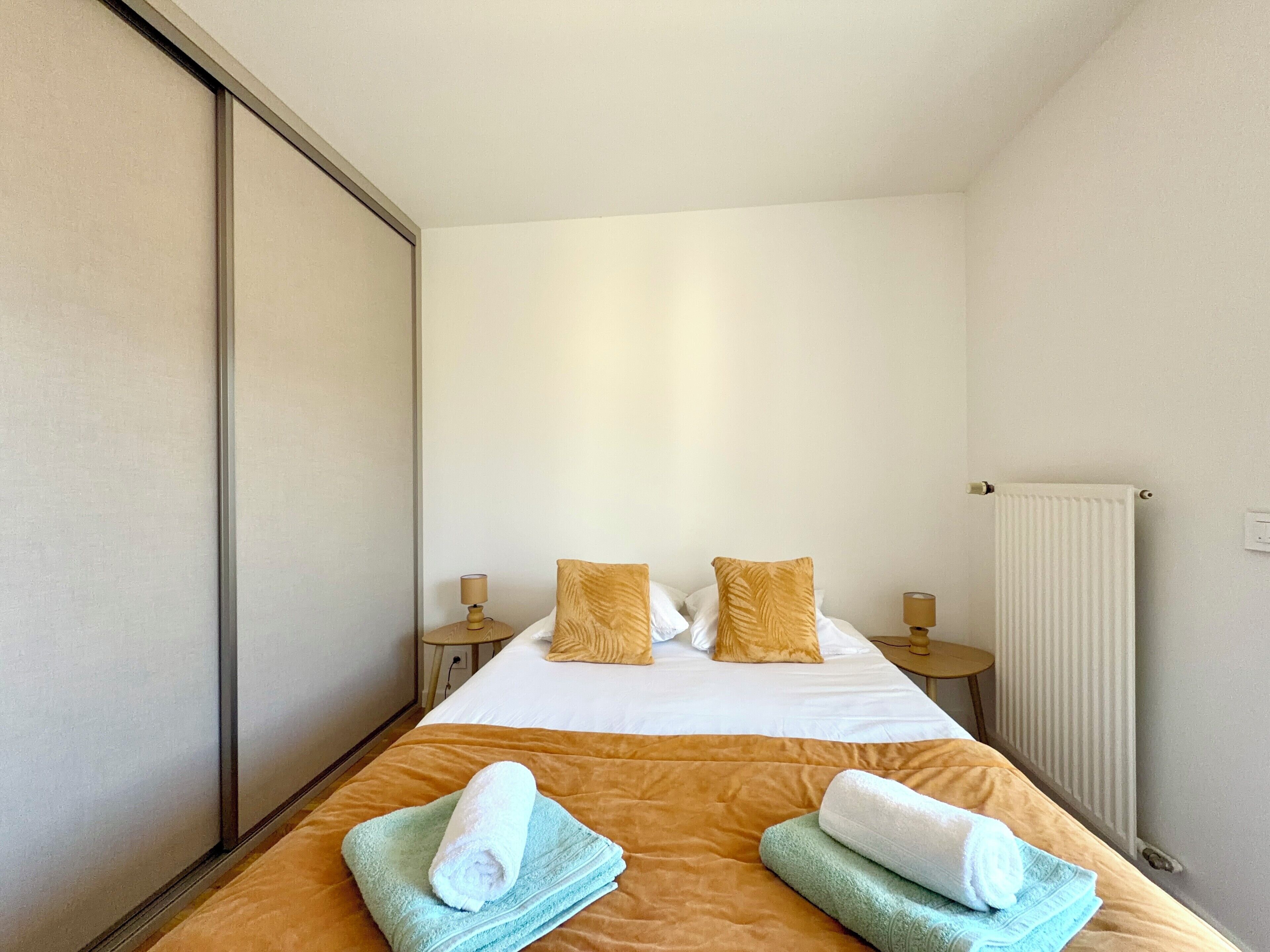 Una habitación de hotel con una cama, dos almohadas, un radiador y dos mesitas de noche con lámparas.