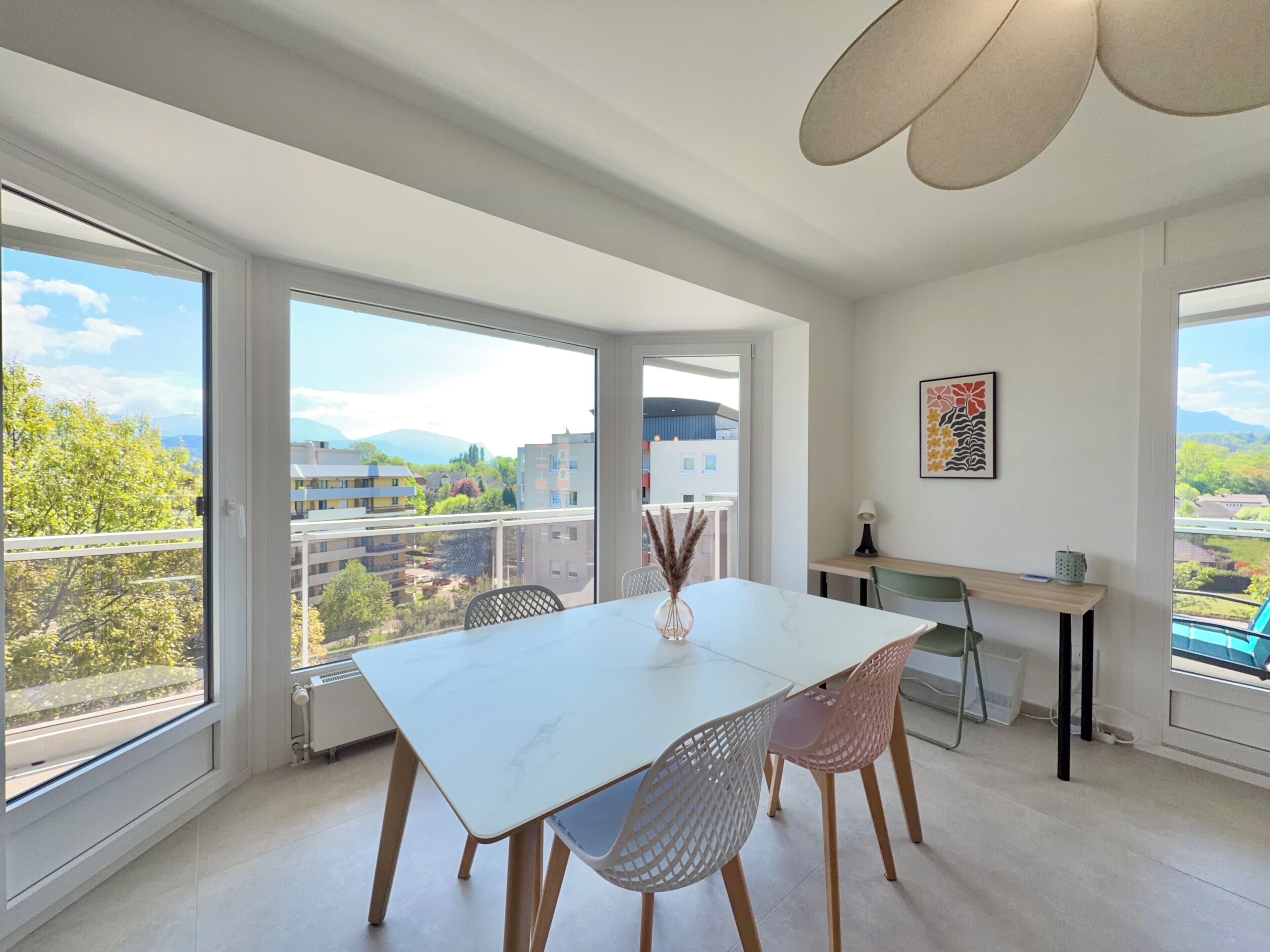 Un espace de repas moderne avec une table et des chaises blanches, de grandes fenêtres offrant une vue sur les bâtiments et la verdure, et un ventilateur de plafond.