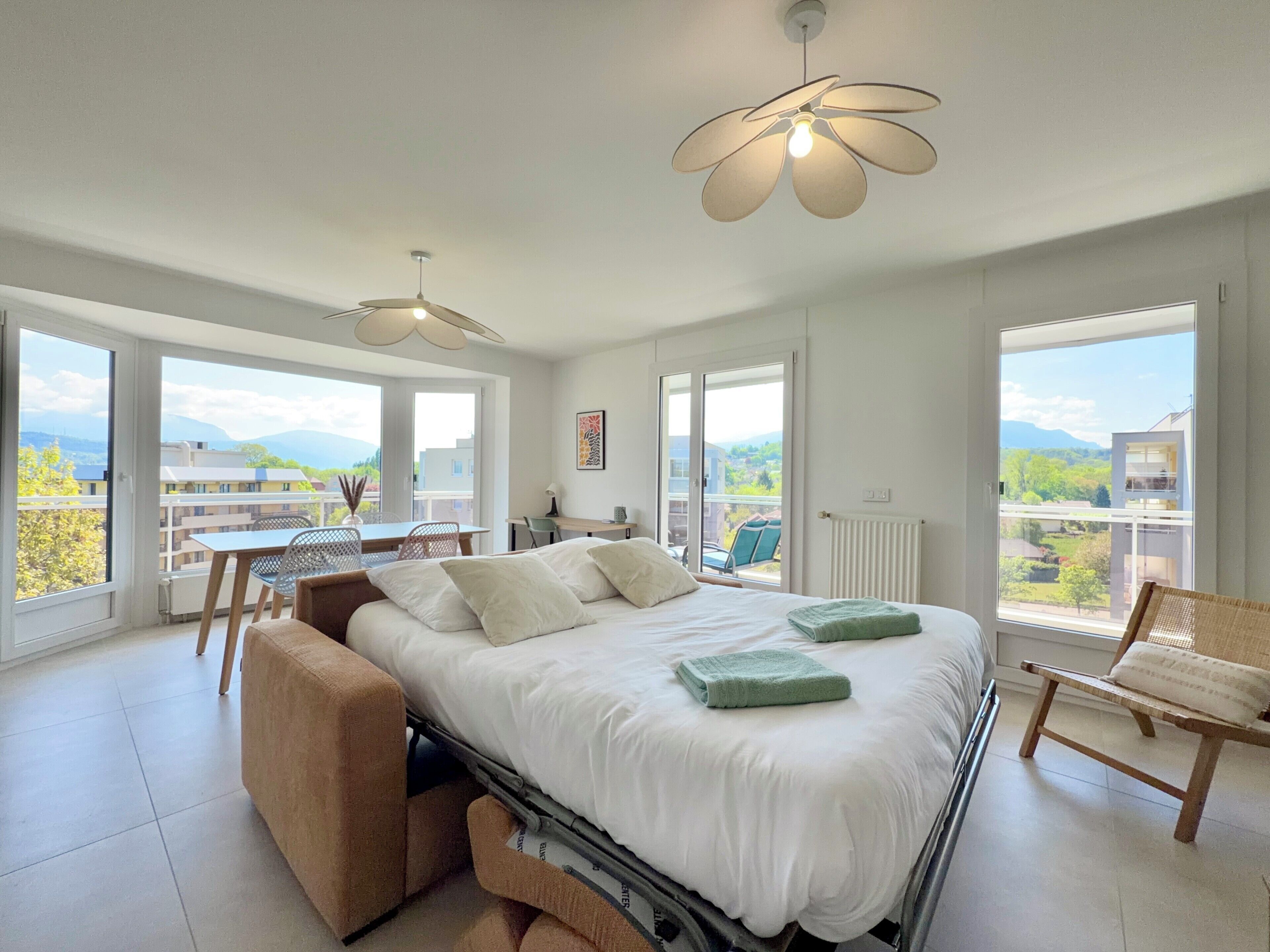 A bedroom with a bed, a chair, a table, and large windows with a view.