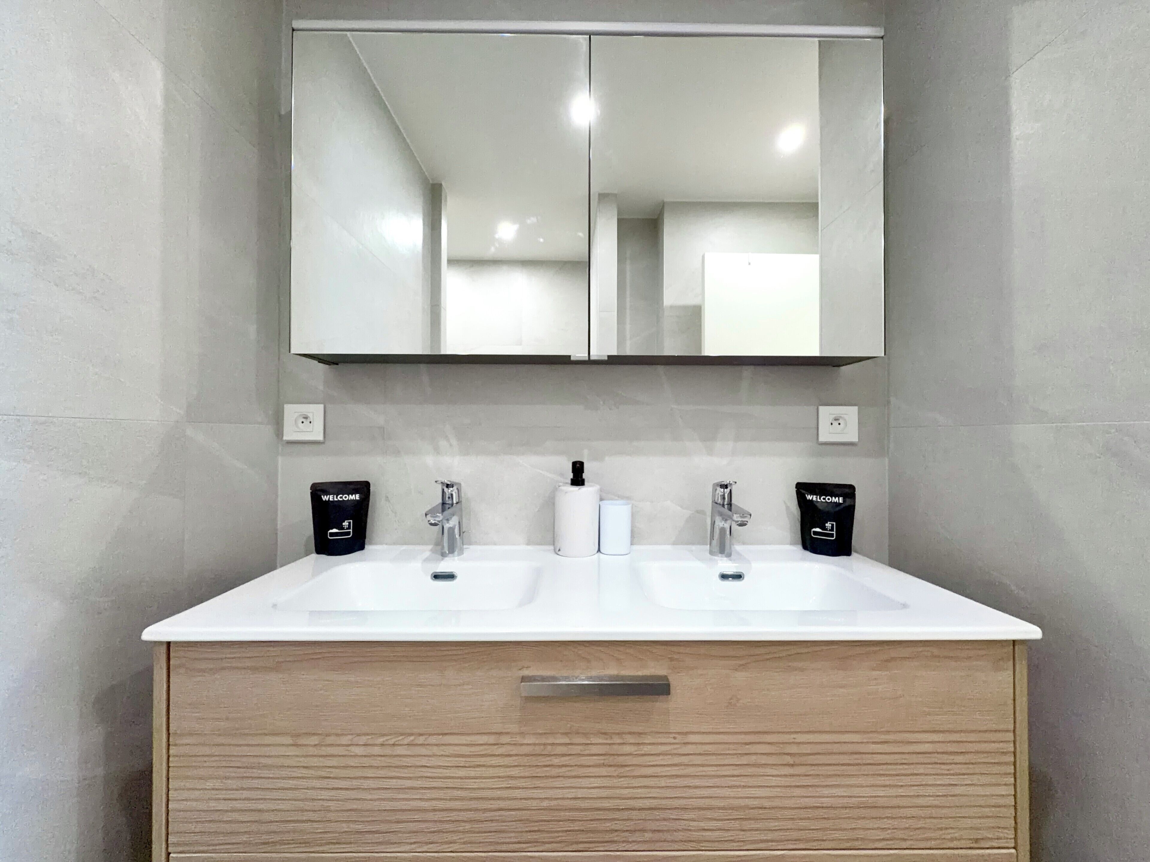 A bathroom with a white sink, two faucets, a mirror, and two black welcome bags on either side.