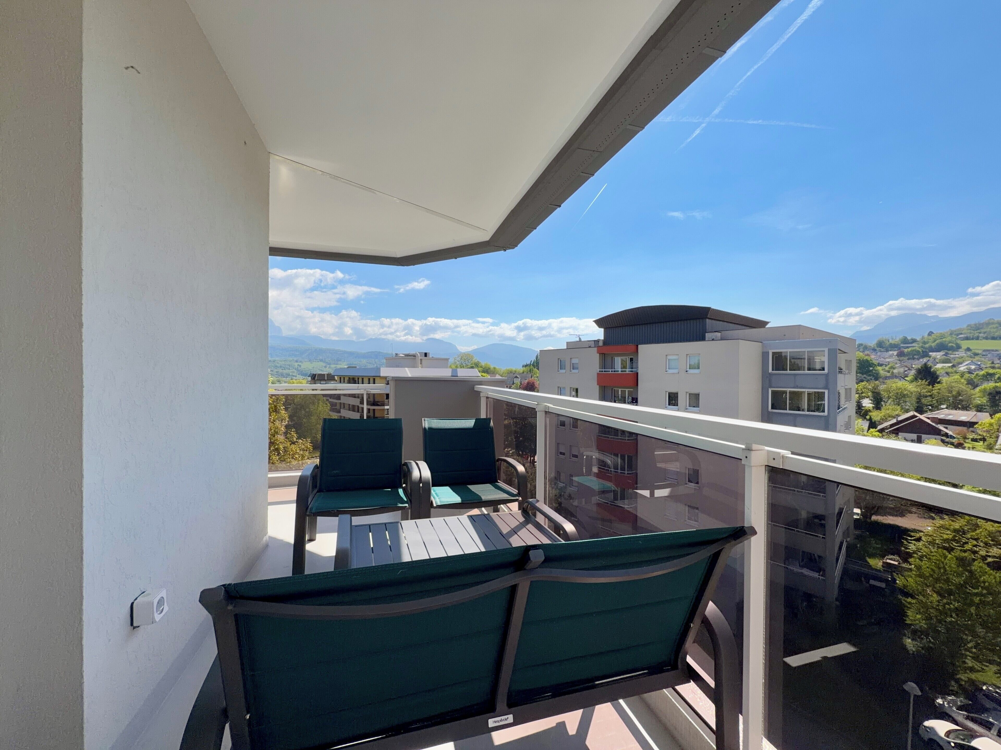 A balcony with two chairs and a table, overlooking a residential area with modern buildings and a clear sky.