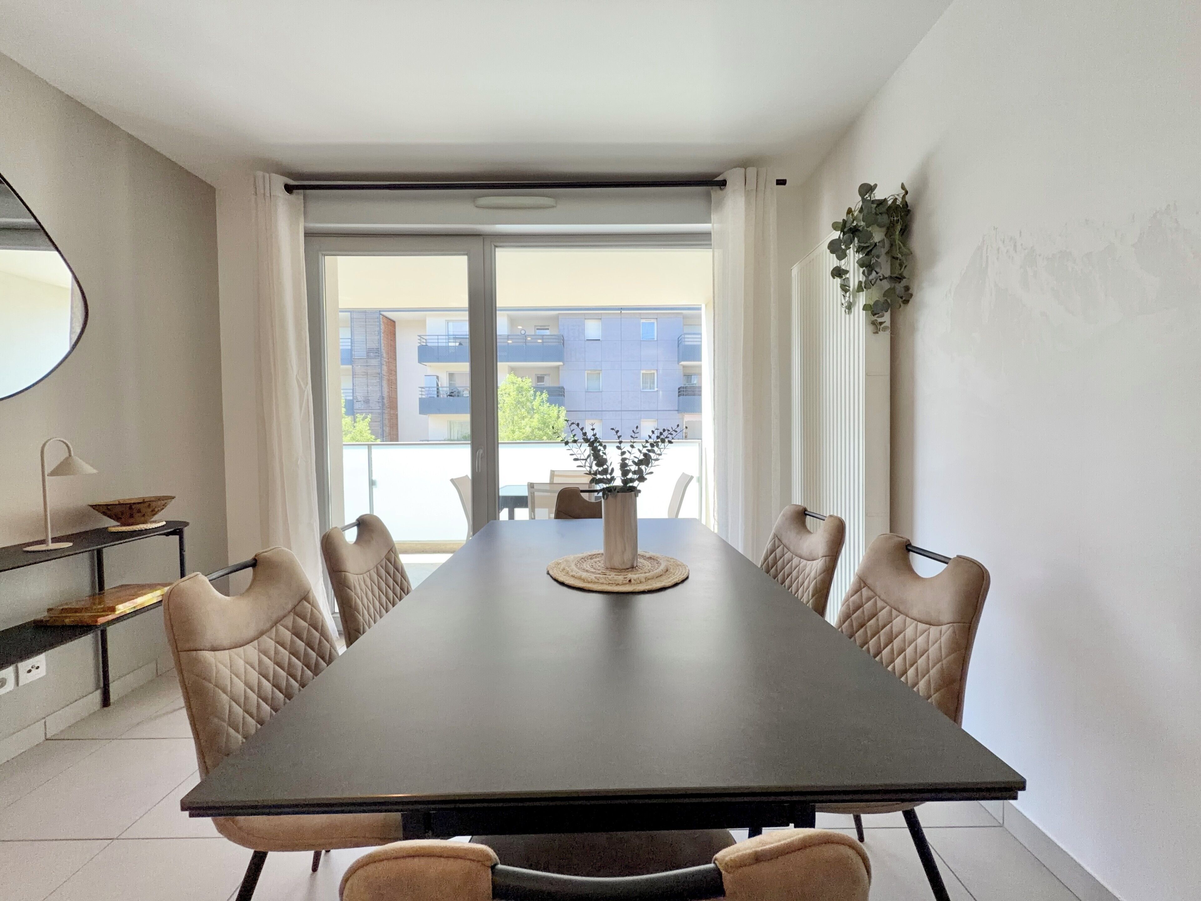 Une salle à manger moderne avec une table sombre, des chaises de couleur beige, un vase avec des plantes et une vue sur les bâtiments à travers de grandes fenêtres.