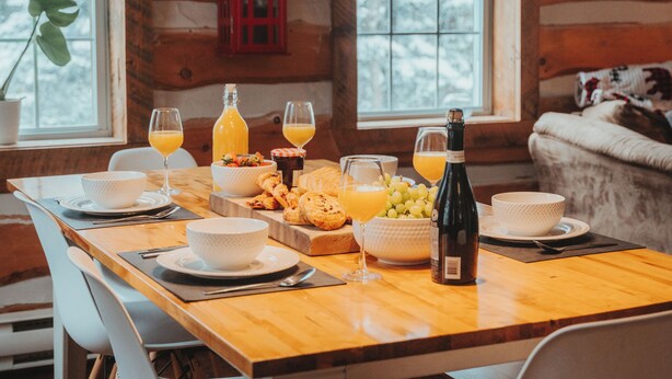 A wooden cabin interior with a dining table set for breakfast, including bowls, glasses, and a bottle of juice. - Authentic Ranch Experience Close to Mont-Tremblant (Val-des-Lacs)