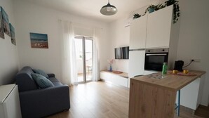 Een modern appartement met een bank, een houten eettafel, een keuken met een televisie en oven, en een glazen schuifdeur naar een balkon.