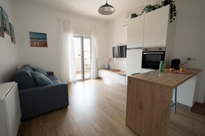 Eine moderne Wohnung mit Sofa, einem Holzesstisch, einer Küche mit Fernseher und Backofen sowie einer Glastür zum Balkon.