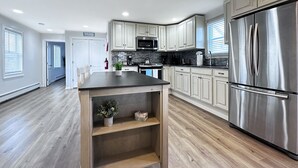 A modern kitchen with white cabinets, a stainless steel refrigerator, and a central island with shelves. - 1BR Apt | High Speed WiFi | Washer/Dryer | Coffee (Seaside Heights)