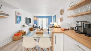 Eine moderne Küche mit Kaffeemaschine, ein Essbereich mit Glastisch und Stühlen sowie ein Wohnbereich mit Sofa und Fernseher.
