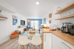 A modern kitchen with a coffee machine, a dining area with a glass table and chairs, and a living area with a sofa and a TV. - Xanadu Retreat - Near Coligny (Hilton Head Island)