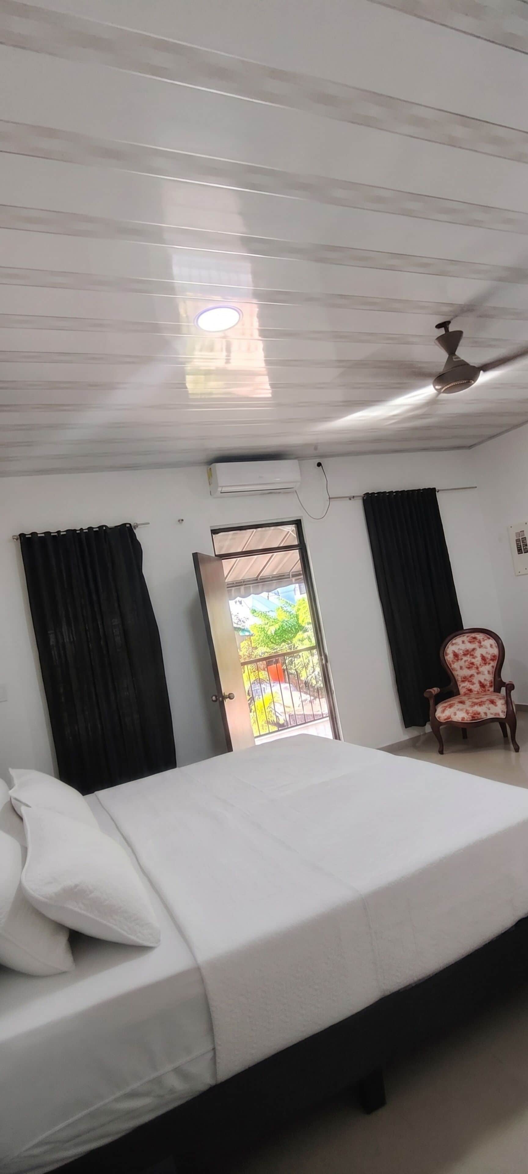 Habitación de hotel con cama, una silla, aire acondicionado y ventilador de techo, con vistas al exterior.