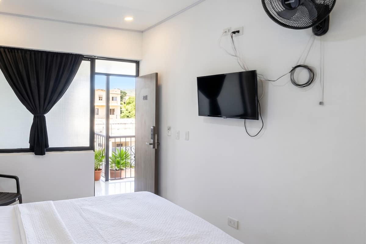 A bedroom with a bed, a chair, a TV, a window with curtains, and a door leading outside.