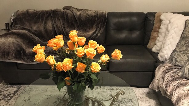 Een glazen salontafel met een vaas vol gele rozen, een donker leren bank en een kamerbreed tapijt met een patroon.