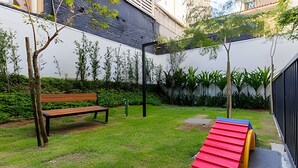 Un espace extérieur verdoyant avec un banc en bois, un toboggan rouge et jaune, et une clôture noire.