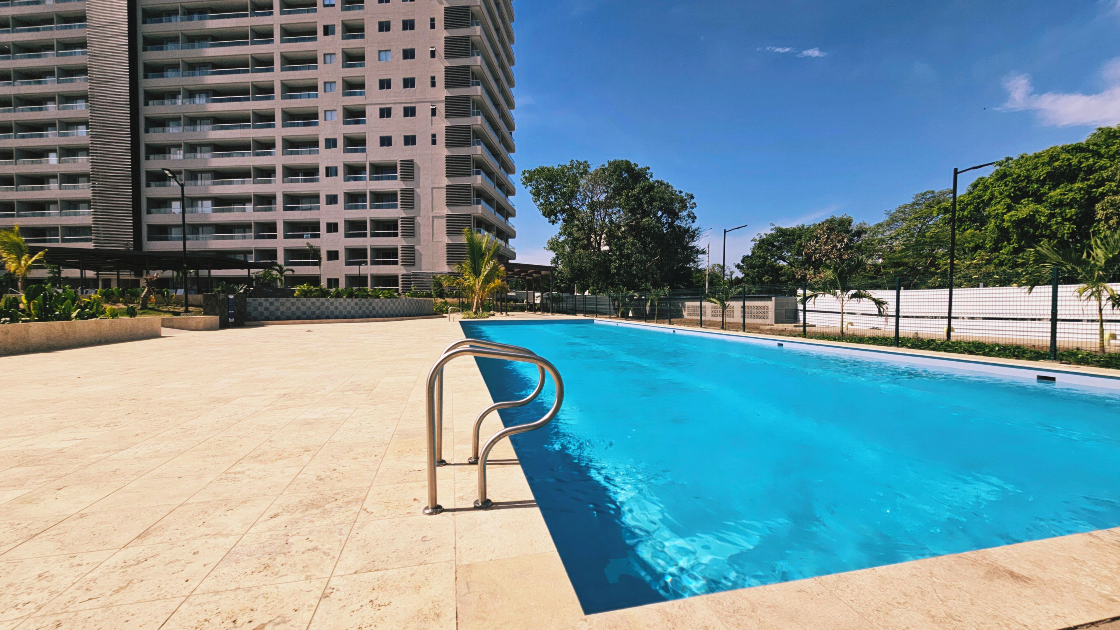 A swimming pool with a metal ladder, surrounded by a tiled deck and a tall building in the background.