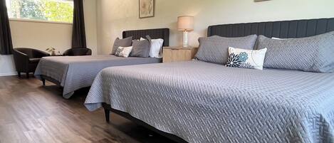A hotel room with two beds, a ceiling fan, a window with curtains, a nightstand with a lamp, and artwork on the wall.
