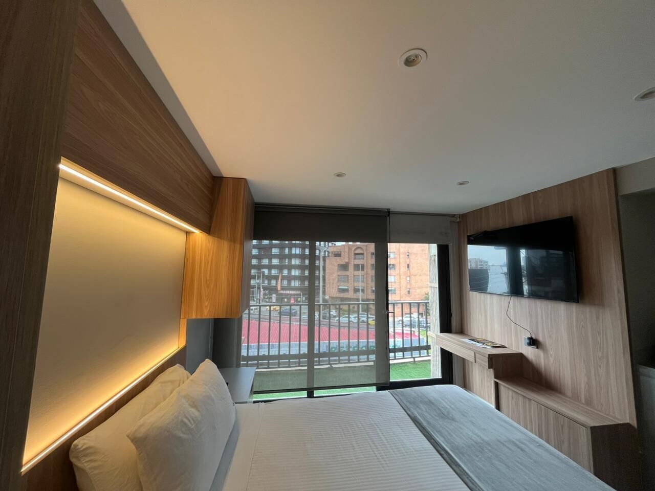 Habitación de hotel moderna con una cama grande, un televisor montado en la pared y vistas a la ciudad por la ventana.