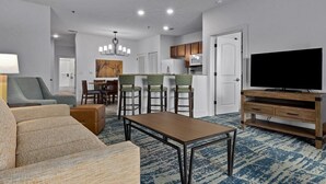 A modern living room with a sofa, a coffee table, a TV, and a dining area with chairs and a table. - Hill Country | Group Nature Retreat | Pool + Trails | 3BR Sleeps 10 (Hunt)