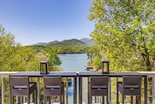 A balcony with wicker chairs and two lanterns overlooking a lake surrounded by trees and hills. - Lake Lure Modern Retreat | Marina Access + Views (Lake Lure)