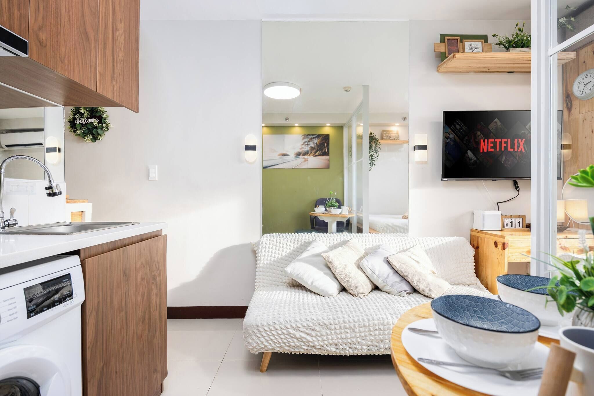 A modern living room with a sofa, a television, a kitchen area with a washing machine, and a dining table with bowls.