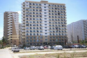 Un edificio residencial de varios pisos con balcones, un estacionamiento con coches y cielo despejado.