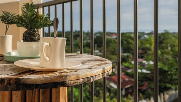 Una mesa de madera con una taza y platillo blancos, una piña y una pequeña planta en maceta en un balcón con barandilla metálica.