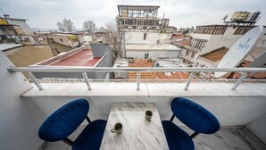 Balcón con una mesa y dos sillas, con vistas a un paisaje urbano que incluye edificios y una zona con tejados rojos.