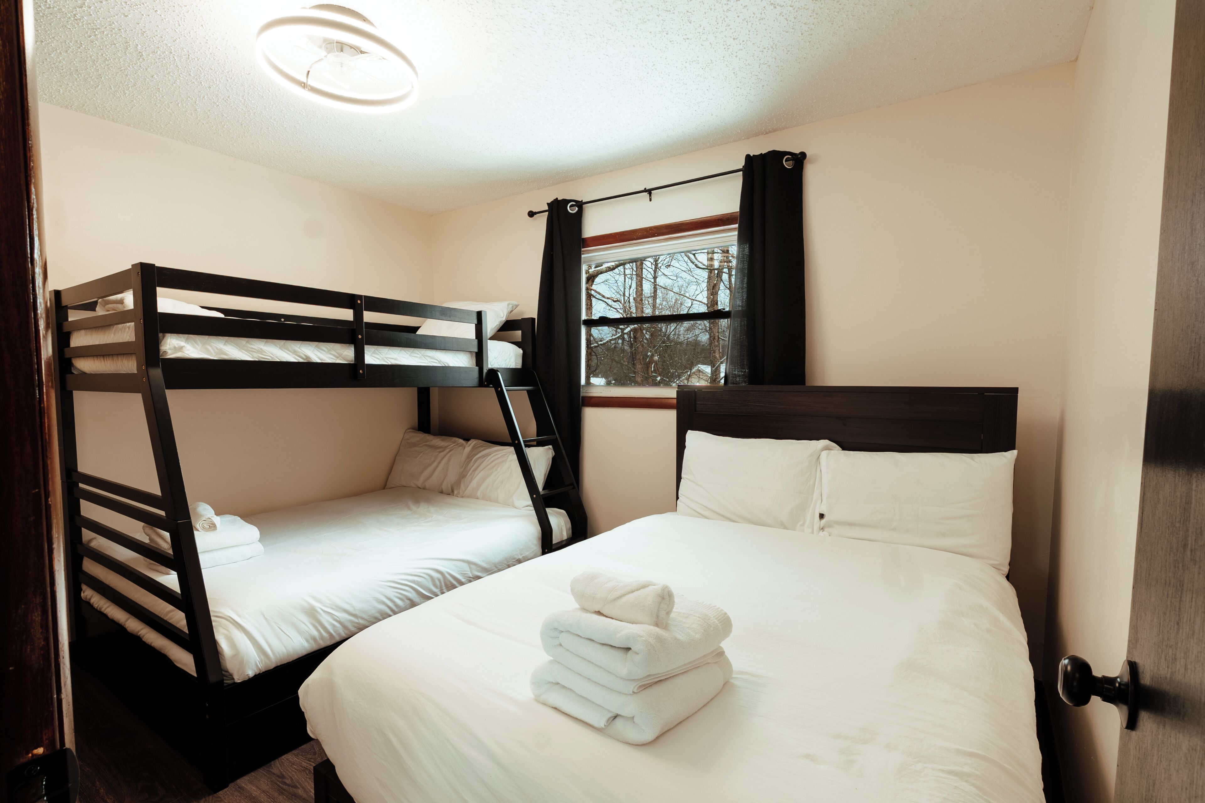 A room with two bunk beds, a window with curtains, a bedside table, and a neatly folded towel on the lower bed.