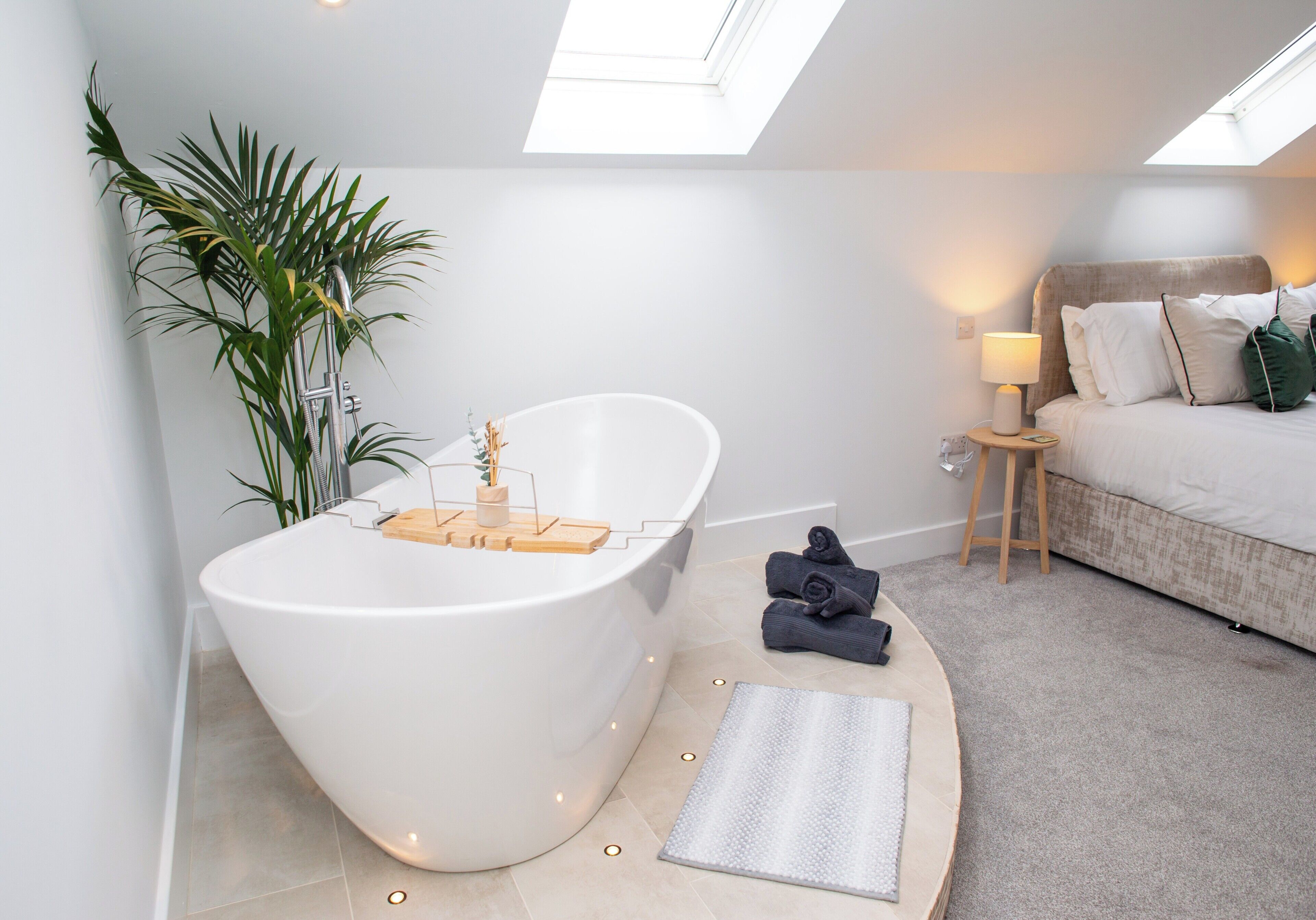 A modern bathroom with a freestanding bathtub, a small bedside table with a lamp, and a bed with pillows.
