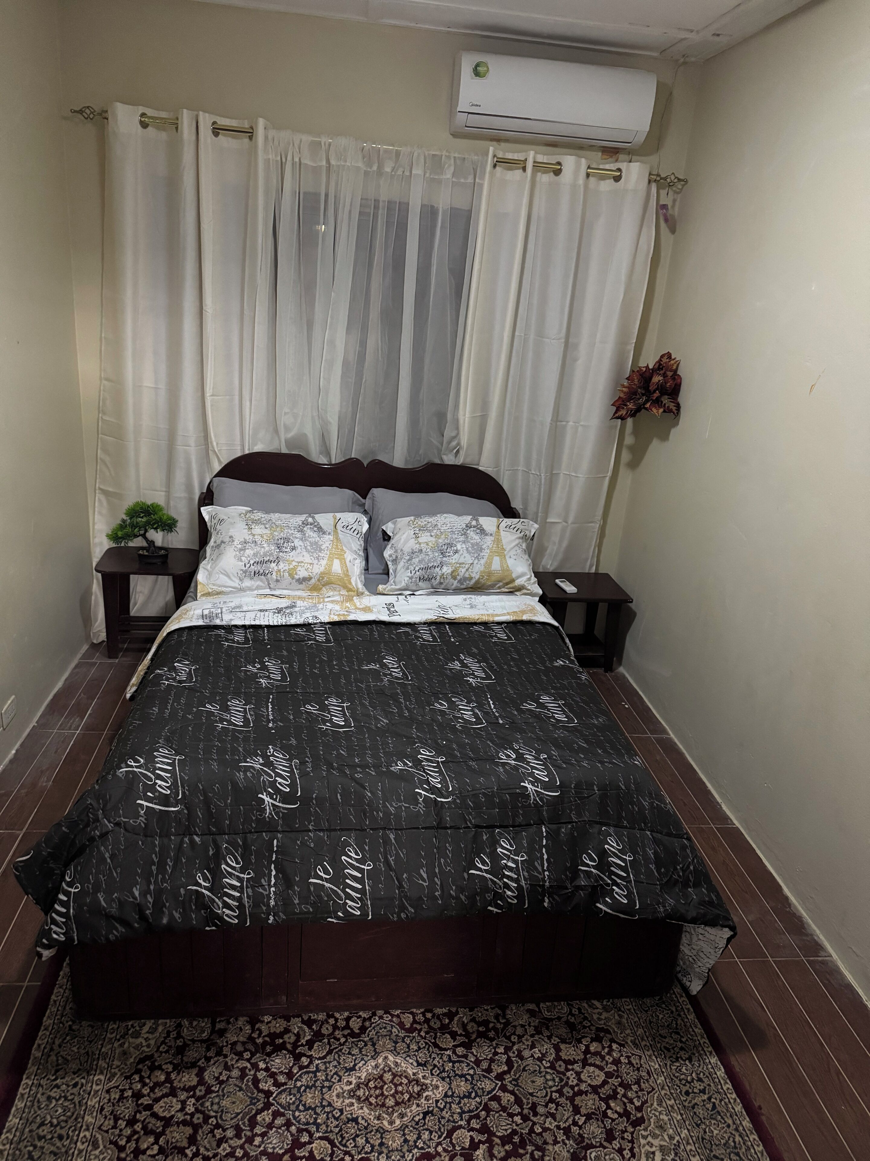 A bedroom with a bed, a small bedside table with a plant, a decorative item on the wall, and an air conditioning unit above the bed.