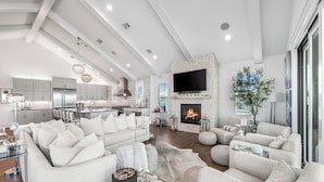 A modern living room with a fireplace, a large flat-screen TV, and a dining area with a kitchen in the background. - Sugar Magnolia: Close to beach in Grayton w/ Pool!  4 Kings + 6 Bunks; sleeps 14 (Santa Rosa Beach)